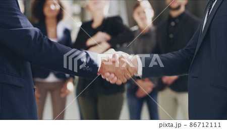 Everyone's ecstatic about this merger. 4k video footage of two businesspeople shaking hands in an office with colleagues applauding in the background. 86712111