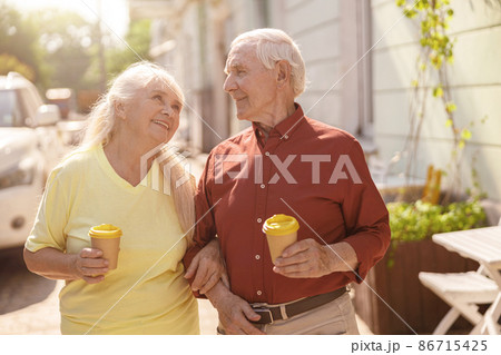 Positive senior family couple with cups walks together along street 86715425