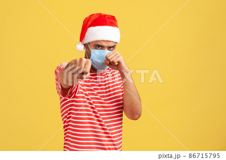 Confident man in santa claus hat with surgical medical mask standing in defensive posture clenched fists, ready to fight with coronavirus covid-19. Indoor studio shot isolated on yellow background 86715795