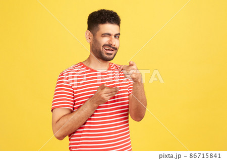 Excited mischievous man with beard in striped t-shirt winking looking and pointing fingers at camera, coquetting and flirting. Indoor studio shot isolated on yellow background Excited mischievous man with beard in striped t-shirt winking looking and pointing fingers at camera, coquetting and flirting. Indoor studio shot isolated on yellow background 86715841
