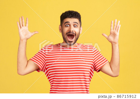 Shocked surprised bearded man in red striped t-shirt looking at camera with big eyes and widely opened mouth, amazed and excited. Indoor studio shot isolated on yellow background 86715912