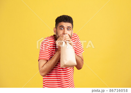 Scared nervous man in striped t-shirt hardly breathing using paper bag and looking at camera with big shocked eyes, trying to calm down. Indoor studio shot isolated on yellow background 86716058