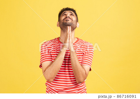 Hopeless desperate man with beard in striped t-shirt folding hands in pray looking up, communicating with god, asking for forgiveness and blessing. Indoor studio shot isolated on yellow background Hopeless desperate man with beard in striped t-shirt folding hands in pray looking up, communicating with god, asking for forgiveness and blessing. Indoor studio shot isolated on yellow background 86716091