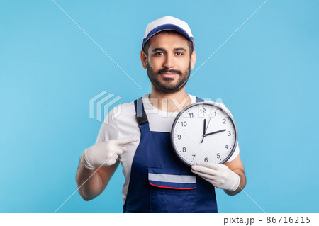 Courier delivery on time. Optimistic positive bearded handyman in overalls and gloves pointing clock, smiling to camera. Professional and punctual service of house repair, maintenance. studio shot 86716215