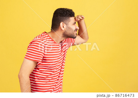 Profile portrait curious bearded man in red striped t-shirt trying to look far away holding hand on forehead, eye vision problems. Indoor studio shot isolated on yellow background 86716473