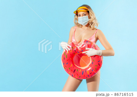 Woman in swimsuit holding rubber ring, wearing hygienic face mask and surgical gloves to prevent contagious coronavirus on resort beach, rest and summer vacation with covid-19. studio shot isolated Woman in swimsuit holding rubber ring, wearing hygienic face mask and surgical gloves to prevent contagious coronavirus on resort beach, rest and summer vacation with covid-19. studio shot isolated 86716515
