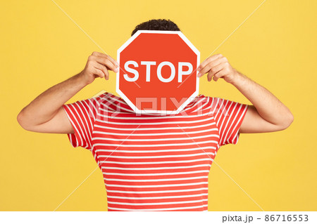 Unknown anonymous man in red striped t-shirt hiding his face behind red stop sign board, scared victim of humiliation and violence. Indoor studio shot isolated on yellow background 86716553