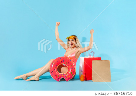 Portrait of enthusiastic overjoyed girl in swimsuit sitting with rubber ring and shopping bags, screaming from happiness, excited by summer sales. indoor studio shot isolated on blue background Portrait of enthusiastic overjoyed girl in swimsuit sitting with rubber ring and shopping bags, screaming from happiness, excited by summer sales. indoor studio shot isolated on blue background 86716710