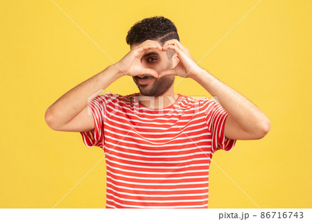 Romantic enamored man with beard in striped t-shirt looking inside heart he made with fingers, showing his feelings, love confession. Indoor studio shot isolated on yellow background 86716743