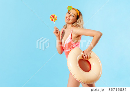 Portrait of lovely carefree beautiful woman in swimsuit holding rubber ring and sweet lollipop candy, enjoying rest on tropical beach, vacation resort. indoor studio shot isolated on blue background Portrait of lovely carefree beautiful woman in swimsuit holding rubber ring and sweet lollipop candy, enjoying rest on tropical beach, vacation resort. indoor studio shot isolated on blue background 86716885