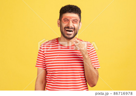 Extremely happy excited man with beard in striped t-shirt holding mask on stick hear eyes pretending he is in eyeglasses, having fun 86716978