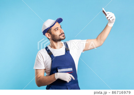 Workman in overalls and gloves talking on video call, communicating online with client using mobile phone, accepting order as delivery, repair and maintenance services. indoor studio shot isolated Workman in overalls and gloves talking on video call, communicating online with client using mobile phone, accepting order as delivery, repair and maintenance services. indoor studio shot isolated 86716979