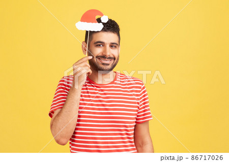 Positive smiling man with beard in red striped t-shirt holding mask on stick with santa claus hat, preparing for christmas and new year celebration. Indoor studio shot isolated on yellow background Positive smiling man with beard in red striped t-shirt holding mask on stick with santa claus hat, preparing for christmas and new year celebration. Indoor studio shot isolated on yellow background 86717026