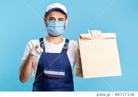 Hey you. Portrait of cheerful bearded handyman in overalls, mask and gloves carrying parcel, pointing to camera. Courier delivering food in paper bag, post mail services. indoor studio shot, isolated Hey you. Portrait of cheerful bearded handyman in overalls, mask and gloves carrying parcel, pointing to camera. Courier delivering food in paper bag, post mail services. indoor studio shot, isolated 86717136