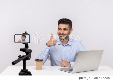 Online webinar, distant training. Elegant successful businessman sitting at workplace, showing thumbs up to mobile phone while recording course, doing blog. studio shot isolated on white background 86717280