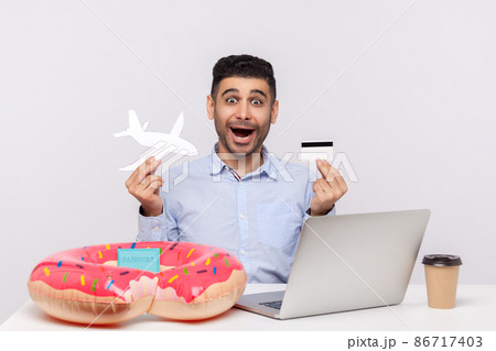 Amazed shocked man holding paper airplane and bank card, sitting in office workplace with rubber ring passport on desk, booking tickets, buying tour, weekend trip. studio shot isolated, travel concept Amazed shocked man holding paper airplane and bank card, sitting in office workplace with rubber ring passport on desk, booking tickets, buying tour, weekend trip. studio shot isolated, travel concept 86717403