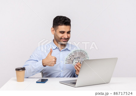 Successful happy businessman sitting in office workplace holding money dollars and showing thumbs up to laptop screen, having online conversation, recommending financial profit. studio shot isolated Successful happy businessman sitting in office workplace holding money dollars and showing thumbs up to laptop screen, having online conversation, recommending financial profit. studio shot isolated 86717524