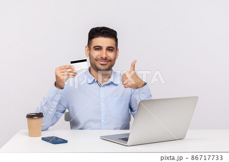 E-commerce. Elegant businessman sitting at workplace with laptop, holding credit card and showing thumbs up, recommending bank service, online purchase. indoor studio shot isolated on white background E-commerce. Elegant businessman sitting at workplace with laptop, holding credit card and showing thumbs up, recommending bank service, online purchase. indoor studio shot isolated on white background 86717733