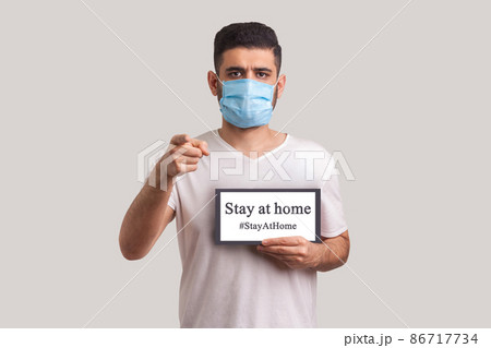 StayAtHome. Man in protective mask holding Stay at home inscription and pointing to camera, warning of coronavirus quarantine, Covid-19 epidemic, preventive measures for spread of infectious disease. StayAtHome. Man in protective mask holding Stay at home inscription and pointing to camera, warning of coronavirus quarantine, Covid-19 epidemic, preventive measures for spread of infectious disease. 86717734