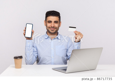Online banking application. Happy businessman sitting in office workplace, holding credit card and mobile phone with empty mock up display, free space for advertisement. indoor studio shot isolated Online banking application. Happy businessman sitting in office workplace, holding credit card and mobile phone with empty mock up display, free space for advertisement. indoor studio shot isolated 86717735