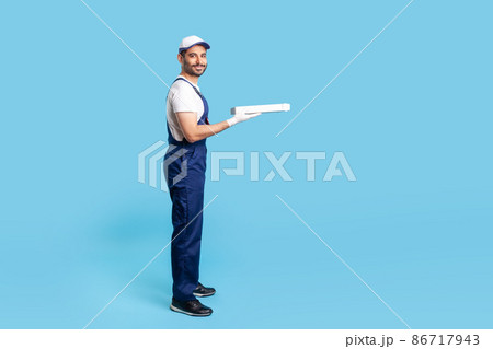 Full length portrait of happy courier in workwear giving pizza box, carrying fast food in protective gloves and smiling to camera. Professional delivery service. indoor studio shot, blue background 86717943