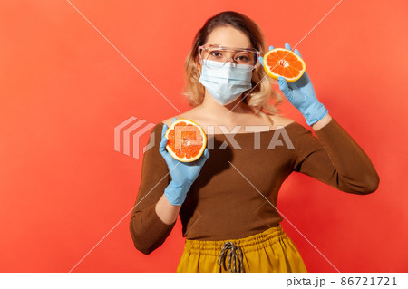 Woman in protective glasses, mask, gloves holding orange fresh fruit, natural vitamins for immune system and flu treatment, healthy nutrition during quarantine. indoor studio shot, red background 86721721