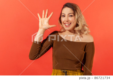 Hello. Portrait of beautiful smiling young lady with blonde hair in brown blouse with raised hand demonstrate hello gesture and ready to make friendship. Indoor studio shoot isolated on red background Hello. Portrait of beautiful smiling young lady with blonde hair in brown blouse with raised hand demonstrate hello gesture and ready to make friendship. Indoor studio shoot isolated on red background 86721734
