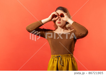 Where you. Portrait of serious blonde woman in casual brown blouse making glasses shape, looking through binoculars gesture. Indoor studio shot isolated on red background. Where you. Portrait of serious blonde woman in casual brown blouse making glasses shape, looking through binoculars gesture. Indoor studio shot isolated on red background. 86721905