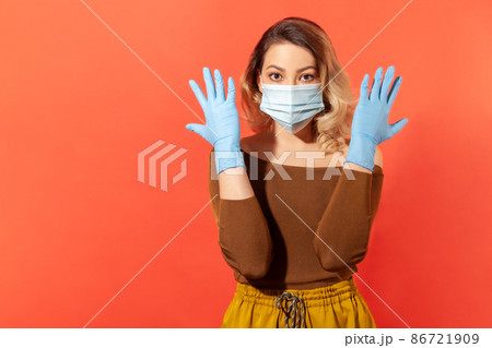 Portrait of woman wearing surgical face mask and gloves to prevent viral disease, coronavirus pandemic threat, 2019-ncov protection recommendations. indoor studio shot isolated on red background Portrait of woman wearing surgical face mask and gloves to prevent viral disease, coronavirus pandemic threat, 2019-ncov protection recommendations. indoor studio shot isolated on red background 86721909