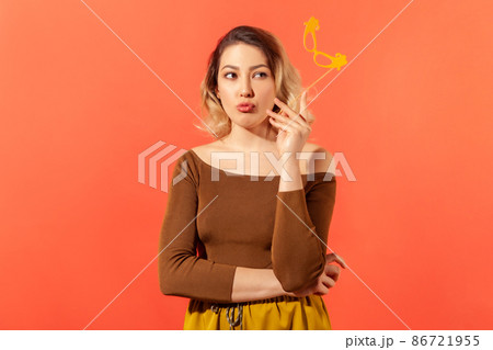 Attractive thoughtful woman holding paper mask on stick contemplating about her costume at fancy party, dreaming. Indoor studio shot isolated on orange background Attractive thoughtful woman holding paper mask on stick contemplating about her costume at fancy party, dreaming. Indoor studio shot isolated on orange background 86721955
