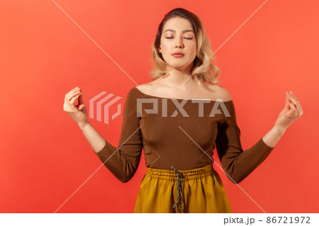 Portrait of beautiful calm young woman with blonde hair in brown blouse standing with closed eyes, meditation with mudra gesture, peaceful mind. Indoor studio shot isolated on red background 86721972