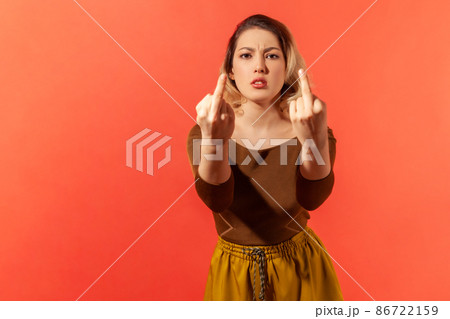 Portrait of angry serious young woman with blonde hair in brown casual blouse standing and shows fuck sign with both hands into the camera. Indoor studio shot isolated on red background 86722159
