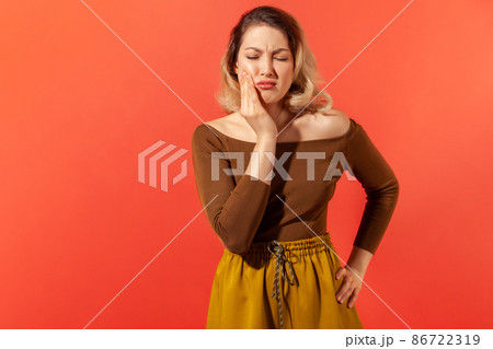Dental problem. Unhealthy sick blonde woman touching cheek, closing eyes with expression of terrible suffer from toothache, sensitive teeth, cavities. Indoor studio shot isolated on red background 86722319