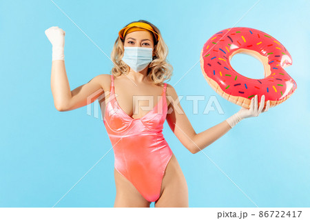Confident woman in swimsuit holding rubber ring, wearing hygienic face mask and surgical gloves to prevent contagious coronavirus on resort beach, raising hand demonstrating power, strength. isolated Confident woman in swimsuit holding rubber ring, wearing hygienic face mask and surgical gloves to prevent contagious coronavirus on resort beach, raising hand demonstrating power, strength. isolated 86722417