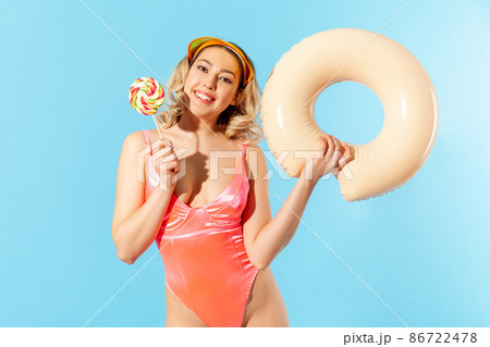 Portrait of lovely carefree beautiful woman in swimsuit holding rubber ring and sweet lollipop candy, enjoying rest on tropical beach, vacation resort. indoor studio shot isolated on blue background Portrait of lovely carefree beautiful woman in swimsuit holding rubber ring and sweet lollipop candy, enjoying rest on tropical beach, vacation resort. indoor studio shot isolated on blue background 86722478