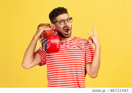 Extremely happy joyful man with beard in eyeglasses and red striped t-shirt holding tape recorder on shoulder and showing rock and roll gesture. Indoor studio shot isolated on yellow background 86722485