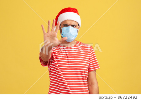 Don't come to me and keep distance. Concerned man in medical face mask and santa claus hat holding hand showing stop gesture. Indoor studio shot isolated on yellow background 86722492