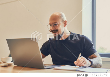 Young caucasian freelancer works with laptop at his desk in office while doing something notes Young caucasian freelancer works with laptop at his desk in office while doing something notes 86725979