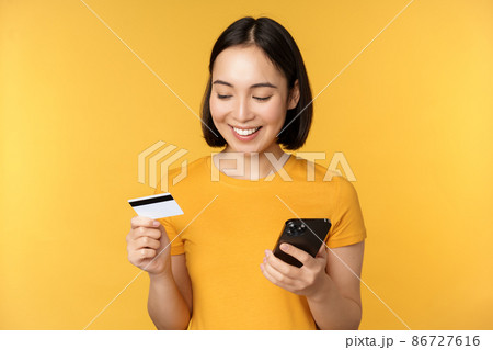 Online shopping. Smiling asian girl using credit card and mobile phone app, paying contactless, order on smartphone application, standing over yellow background 86727616