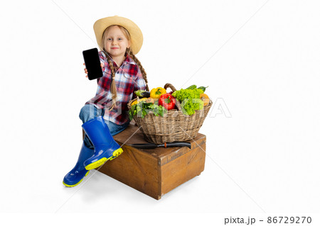Beautiful cute little girl, emotive kid in image of farmer, gardener with large basket of vegetables using phone isolated on white background. 86729270
