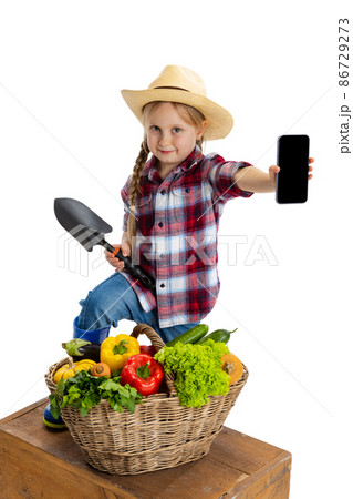 Beautiful cute little girl, emotive kid in image of farmer, gardener with large basket of vegetables using phone isolated on white background. 86729273
