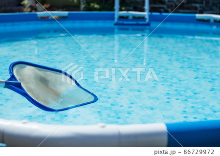 Leaf netting for cleaning leaves from a swimming pool against a backdrop of clear water. Round frame pool and net skimmer in blue water in the backyard of a country house Leaf netting for cleaning leaves from a swimming pool against a backdrop of clear water. Round frame pool and net skimmer in blue water in the backyard of a country house 86729972