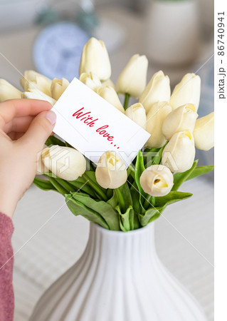 Woman pulling greeting card with love words (text) from bouquet of white tulips flowers in a white vase on the table. Woman pulling greeting card with love words (text) from bouquet of white tulips flowers in a white vase on the table. 86740941