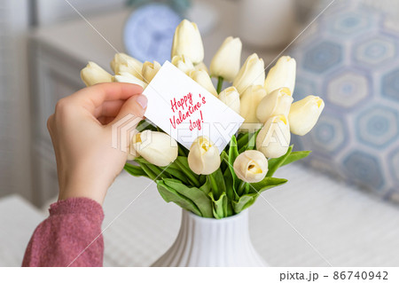 Woman pulling greeting card with Happy Valentine's day words from bouquet of white tulips flowers in a white vase on the table. Woman pulling greeting card with Happy Valentine's day words from bouquet of white tulips flowers in a white vase on the table. 86740942