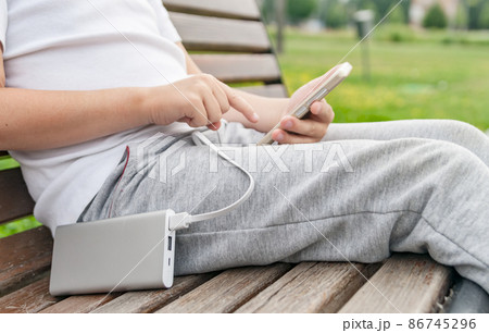 Boy using phone on the bench while charging from the power bank. 86745296