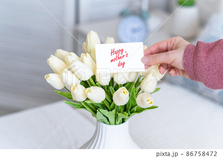 Woman pulling greeting card with Happy mother's day words from bouquet of white tulips flowers in a white vase on the table. 86758472