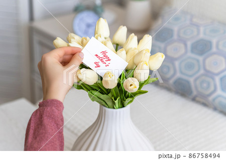 Woman pulling greeting card with Happy mother's day words from bouquet of white tulips flowers in a white vase on the table. 86758494
