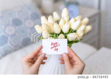 Woman pulling greeting card with Happy mother's day words from bouquet of white tulips flowers in a white vase on the table. 86758495