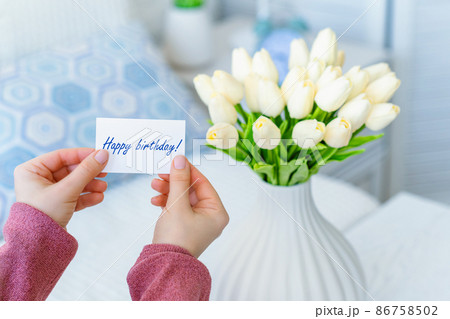 Woman pulling greeting card with Happy birthday words from bouquet of white tulips flowers in a white vase on the table. 86758502