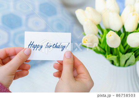 Woman pulling greeting card with Happy birthday words from bouquet of white tulips flowers in a white vase on the table. 86758503